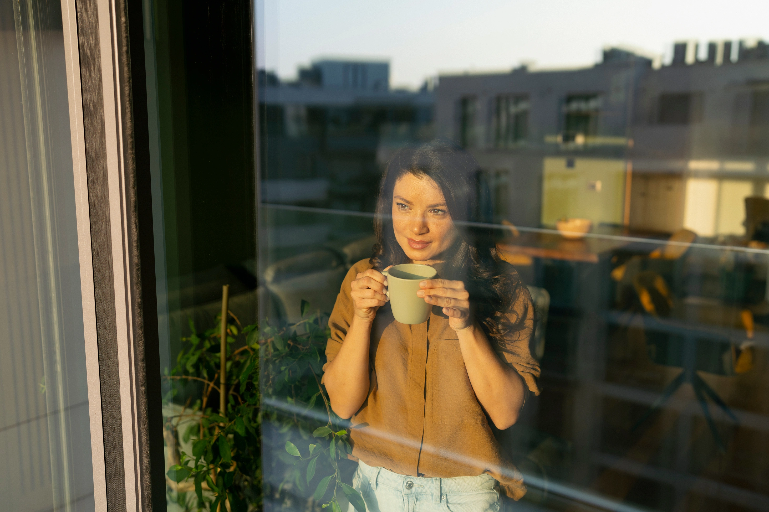 Eine Frau steht mit einer Tasse Kaffee in der Hand am Fenster und blickt hinaus auf die Gebäude in der Ferne. Sonnenlicht fällt herein und sorgt für eine warme Atmosphäre im Raum.