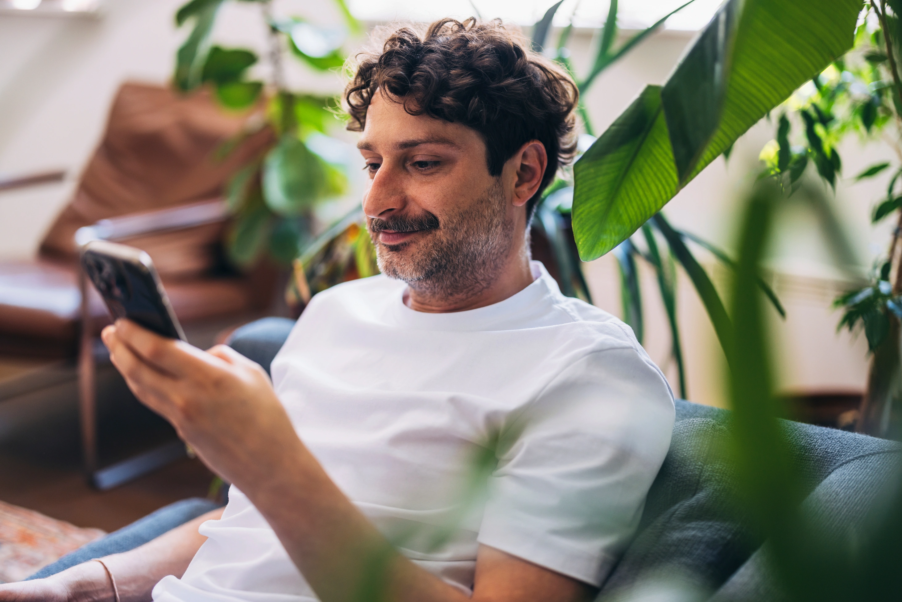 Ein Mann sitzt bequem in seinem Wohnraum, umgeben von sattgrünen Pflanzen, und ist aufmerksam mit seinem Smartphone beschäftigt.