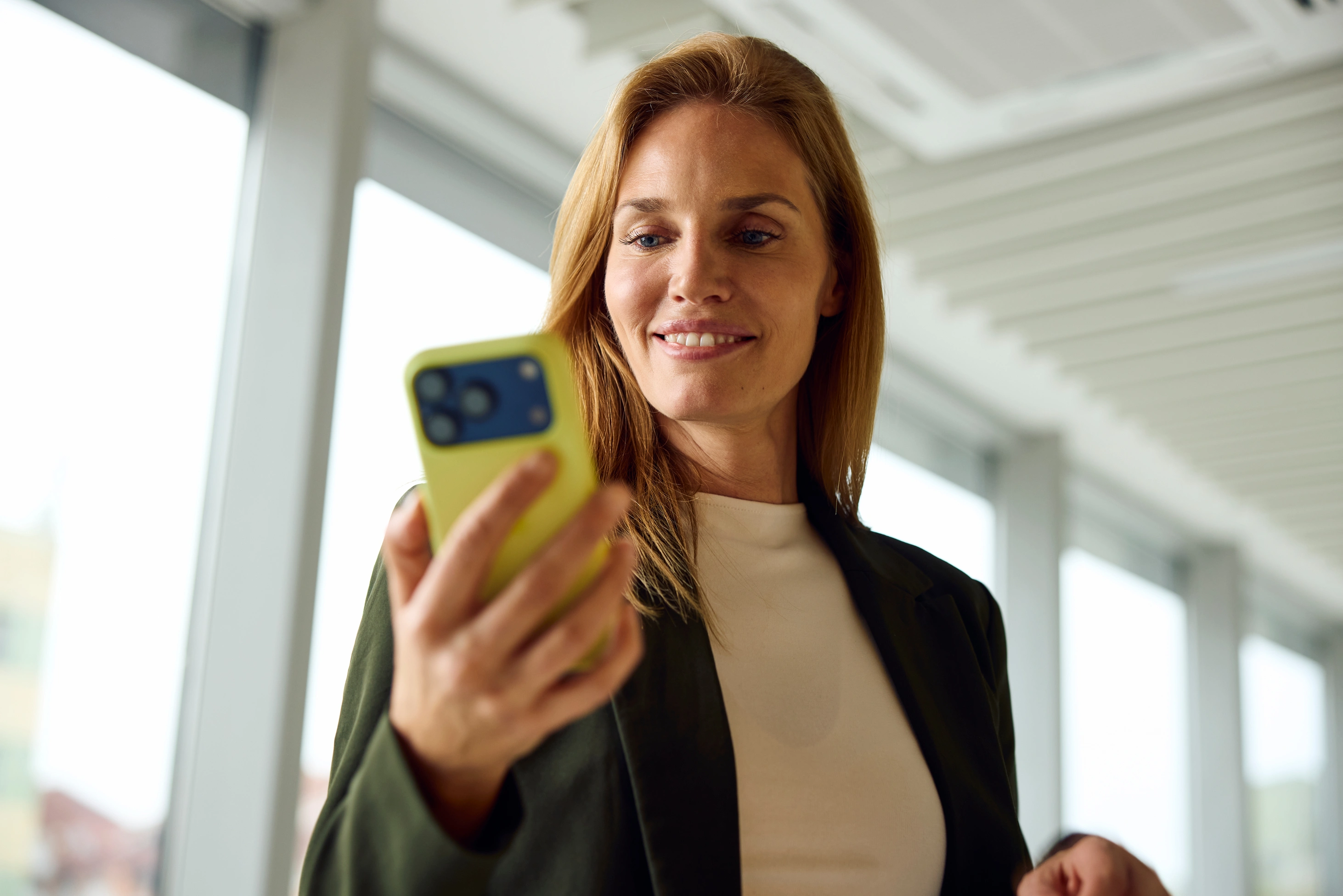 Eine Frau in eleganter Business-Kleidung steht in einem Flur und ist mit ihrem Smartphone beschäftigt.