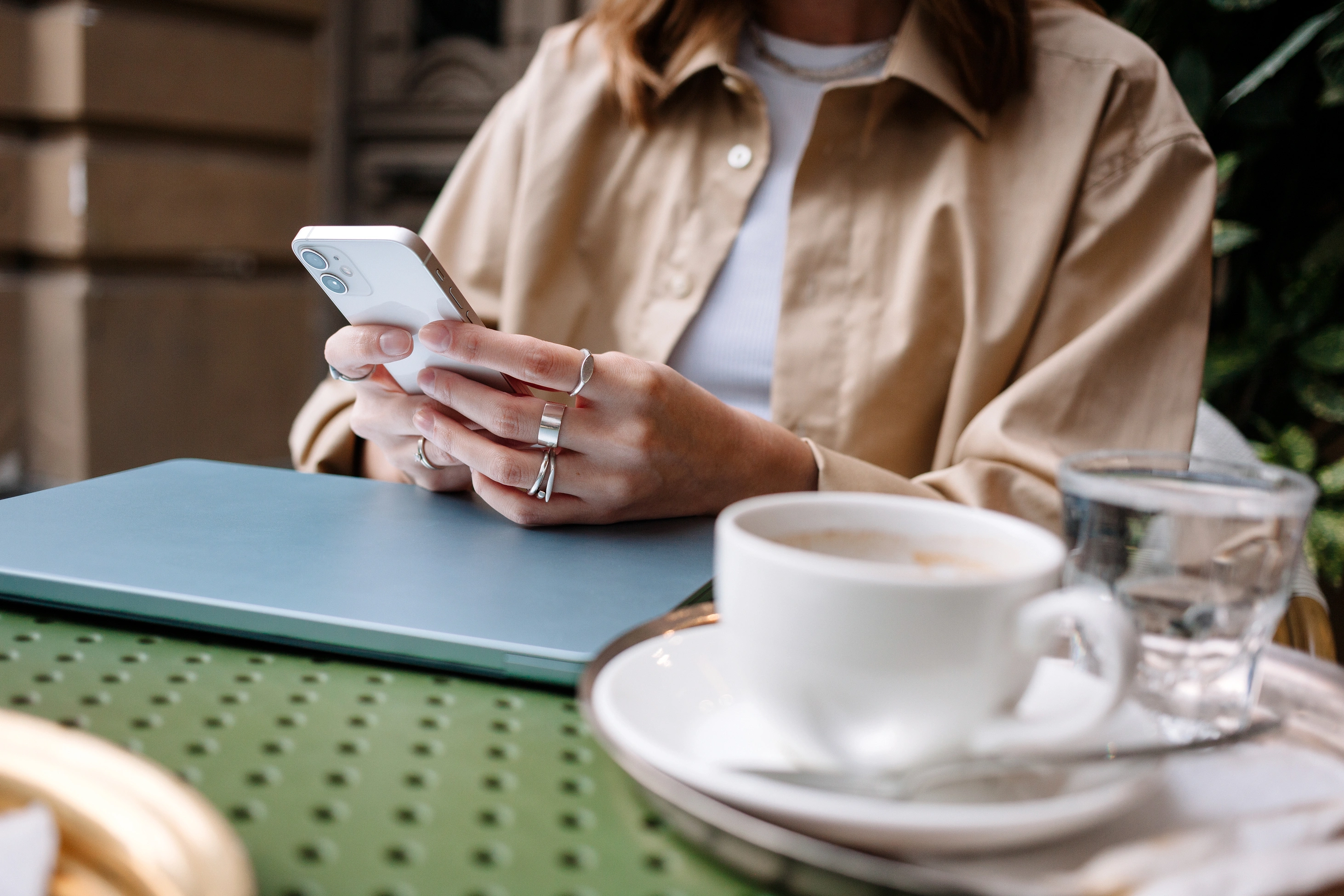 Eine junge Frau sitzt in einem Café an einem grünen Tisch, trinkt Kaffee und scrollt durch ihr Smartphone. Tageslicht erfüllt den gemütlichen Raum und sorgt für eine entspannte Atmosphäre.