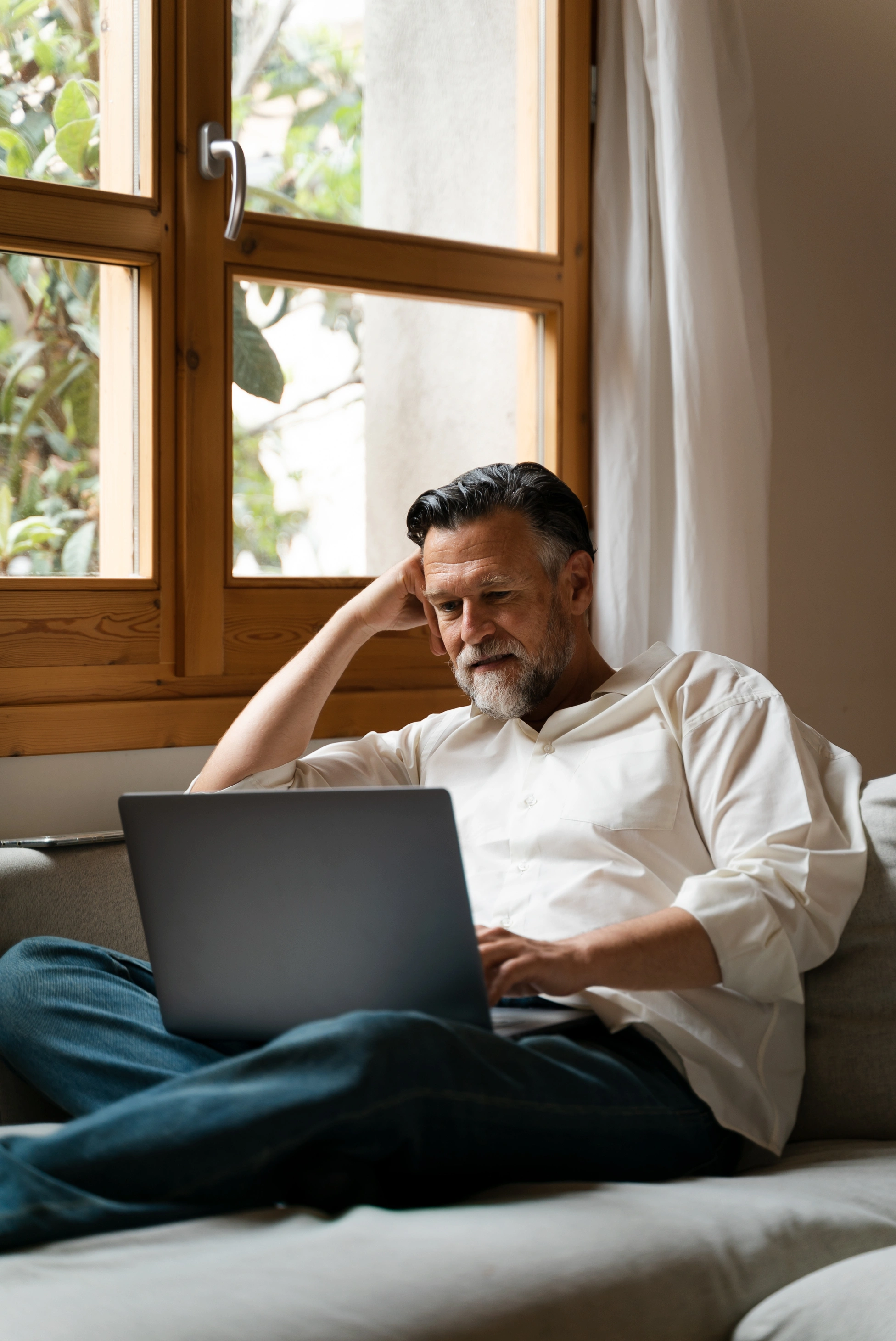 Ein älterer Mann in legerer Kleidung sitzt auf dem Sofa und tippt konzentriert auf seinem Laptop, während er an einem freiberuflichen Projekt arbeitet.