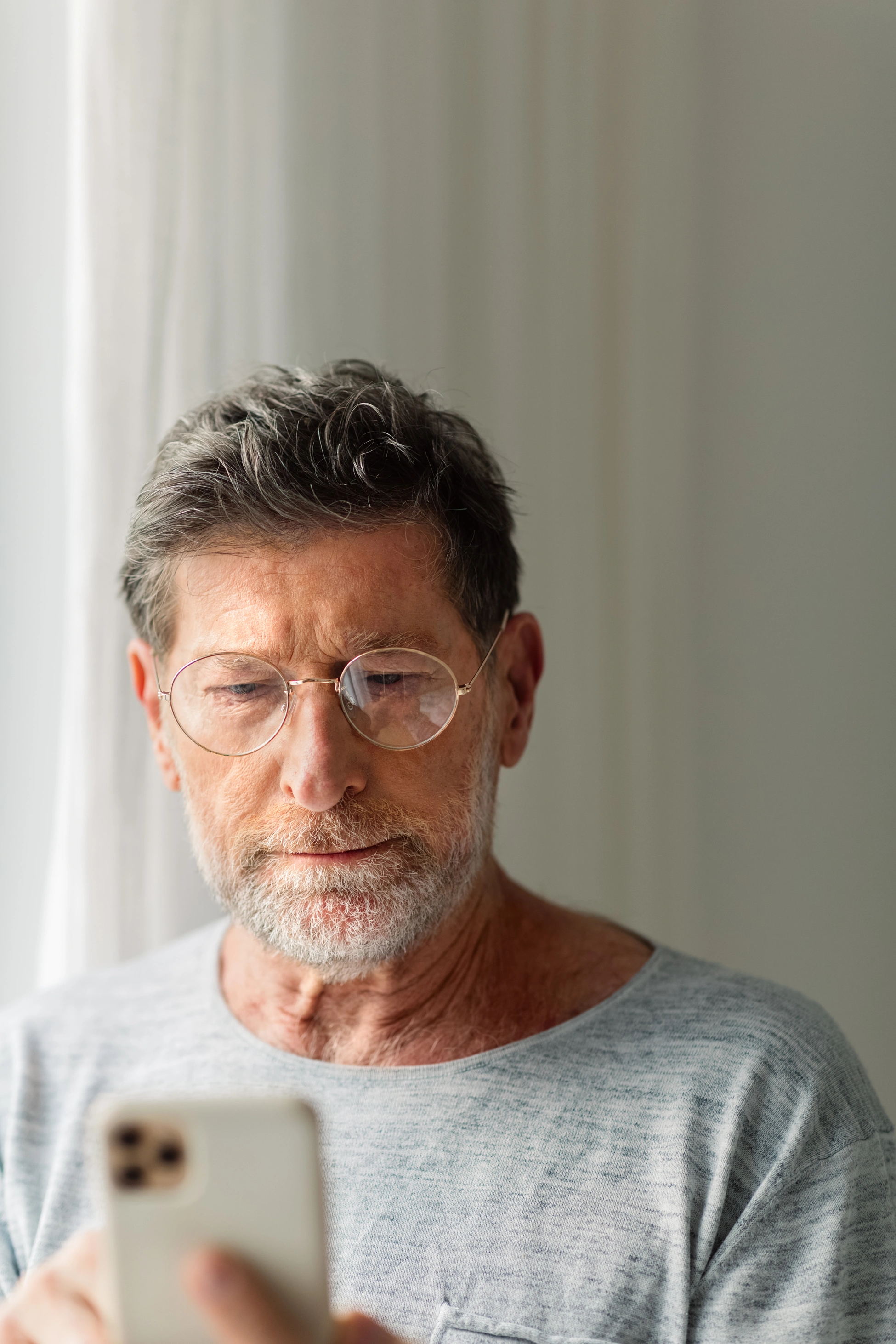 Nahaufnahme eines älteren Mannes mit Brille in legerer Kleidung, der zu Hause sein Smartphone nutzt.