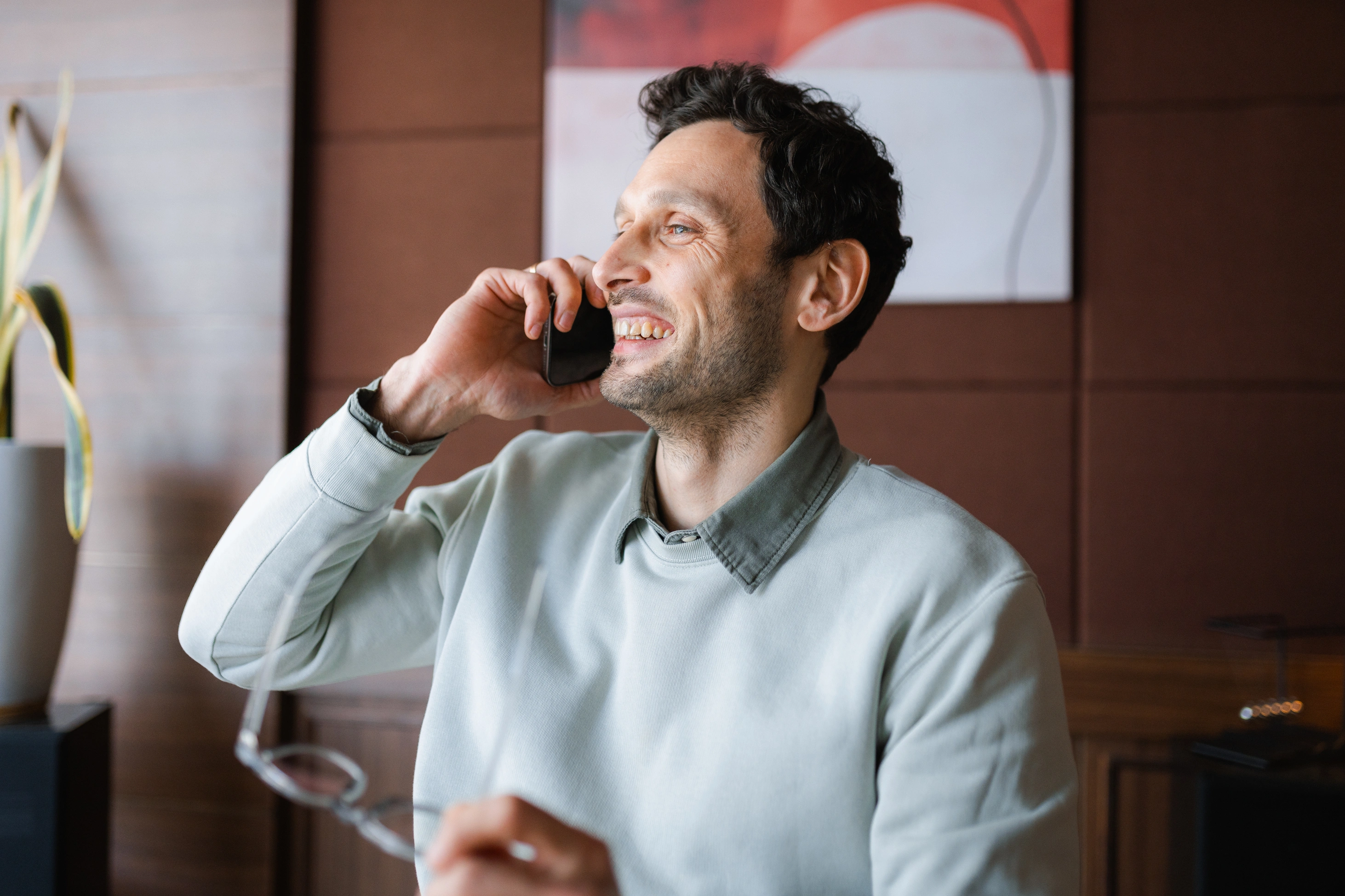 Ein Mann sitzt bei Tageslicht an seinem Schreibtisch im Büro und telefoniert lächelnd mit seinem Smartphone.