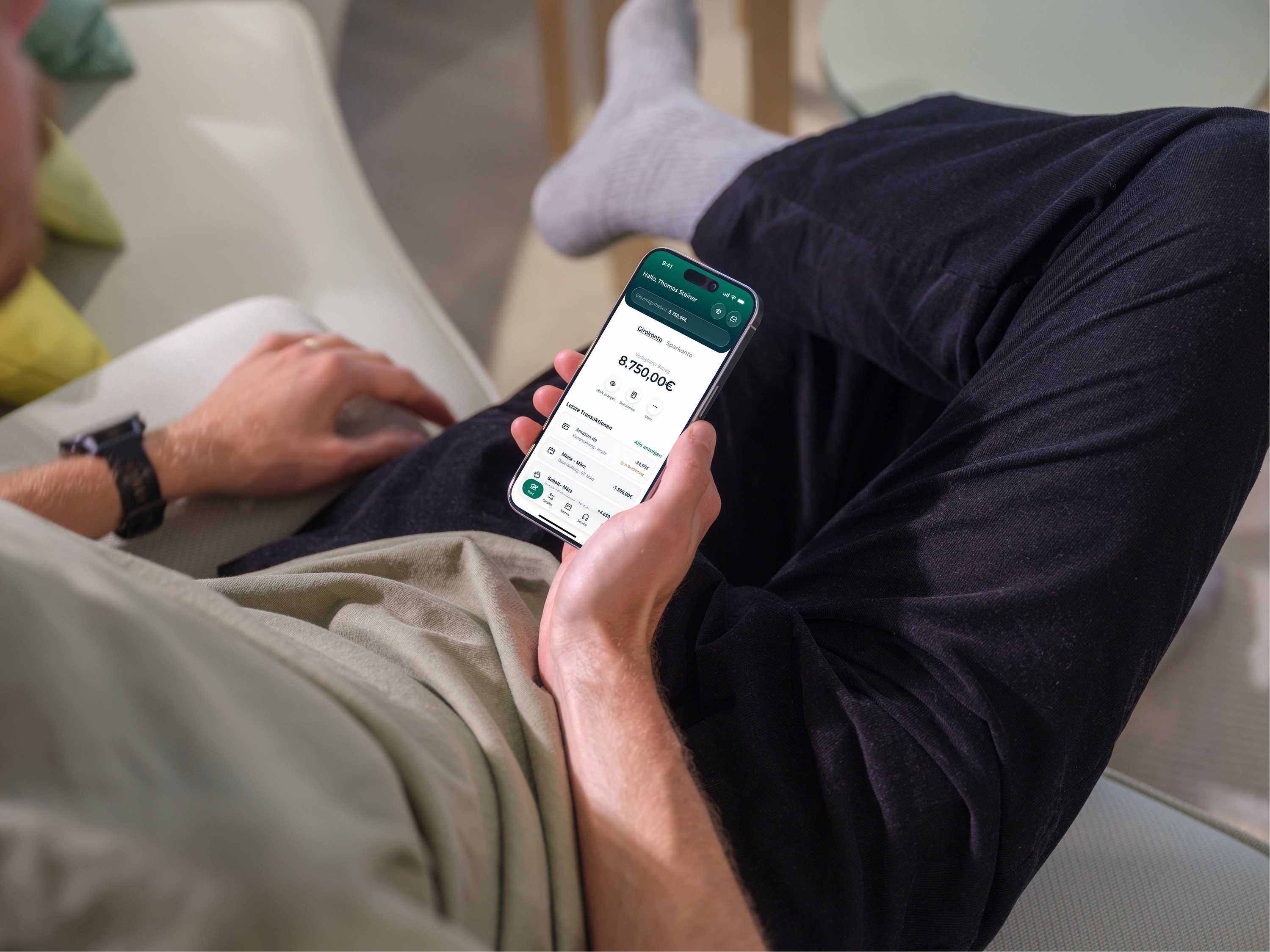 Person sitzt entspannt auf einem Sofa und hält ein Smartphone mit geöffneter Crédit Agricole Banking-App und angezeigtem Kontostand in der Hand.