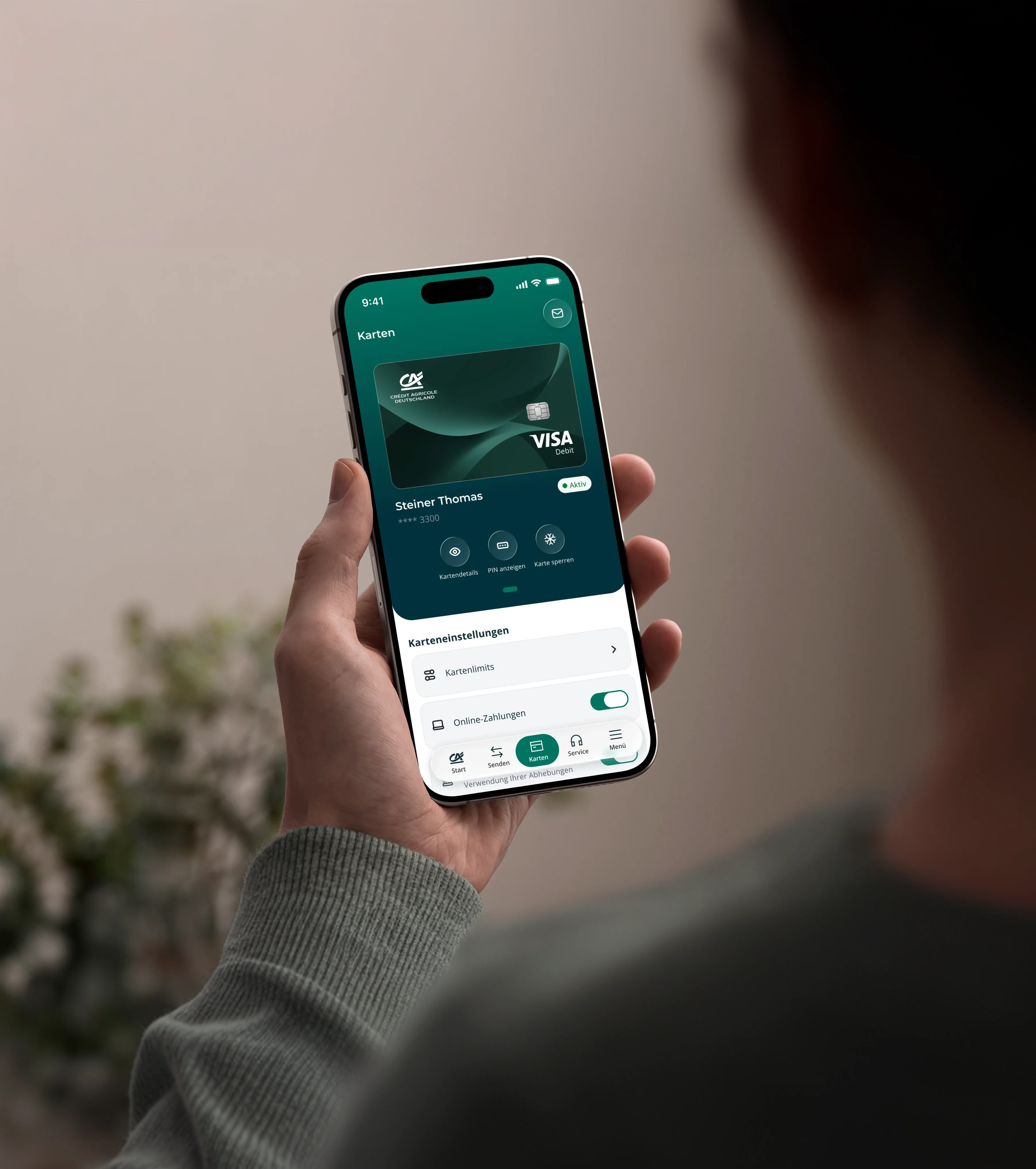 Eine Person hält ein Smartphone mit geöffneter Banking-App. Auf dem Bildschirm ist eine Visa-Debitkarte mit Kartendetails und Karteneinstellungen zu sehen, zum Beispiel Kartenlimits und Online-Zahlungen. Im unscharfen Hintergrund stehen eine helle Wand und eine Grünpflanze.