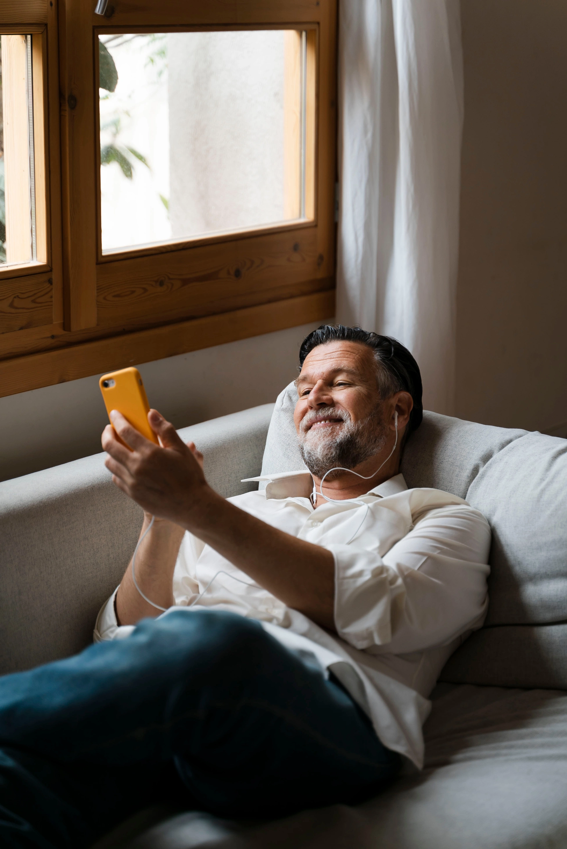 Ein entspannter Mann mittleren Alters liegt zu Hause auf dem Sofa, nutzt sein Smartphone und hört Musik über Kopfhörer.