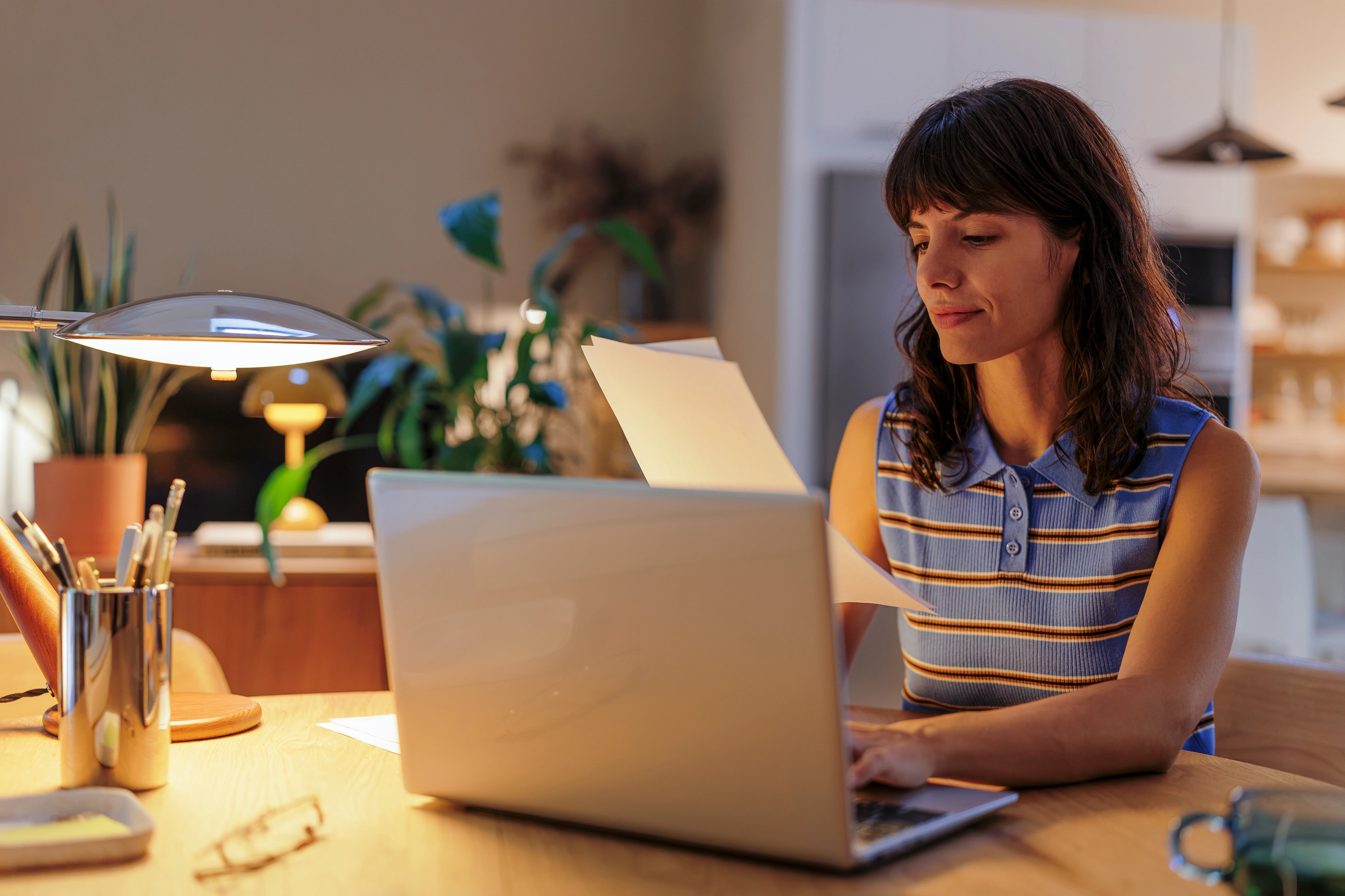 Eine brünette Frau arbeitet in gemütlicher Abendstimmung konzentriert im Homeoffice, tippt auf ihrem Laptop und geht Unterlagen durch.