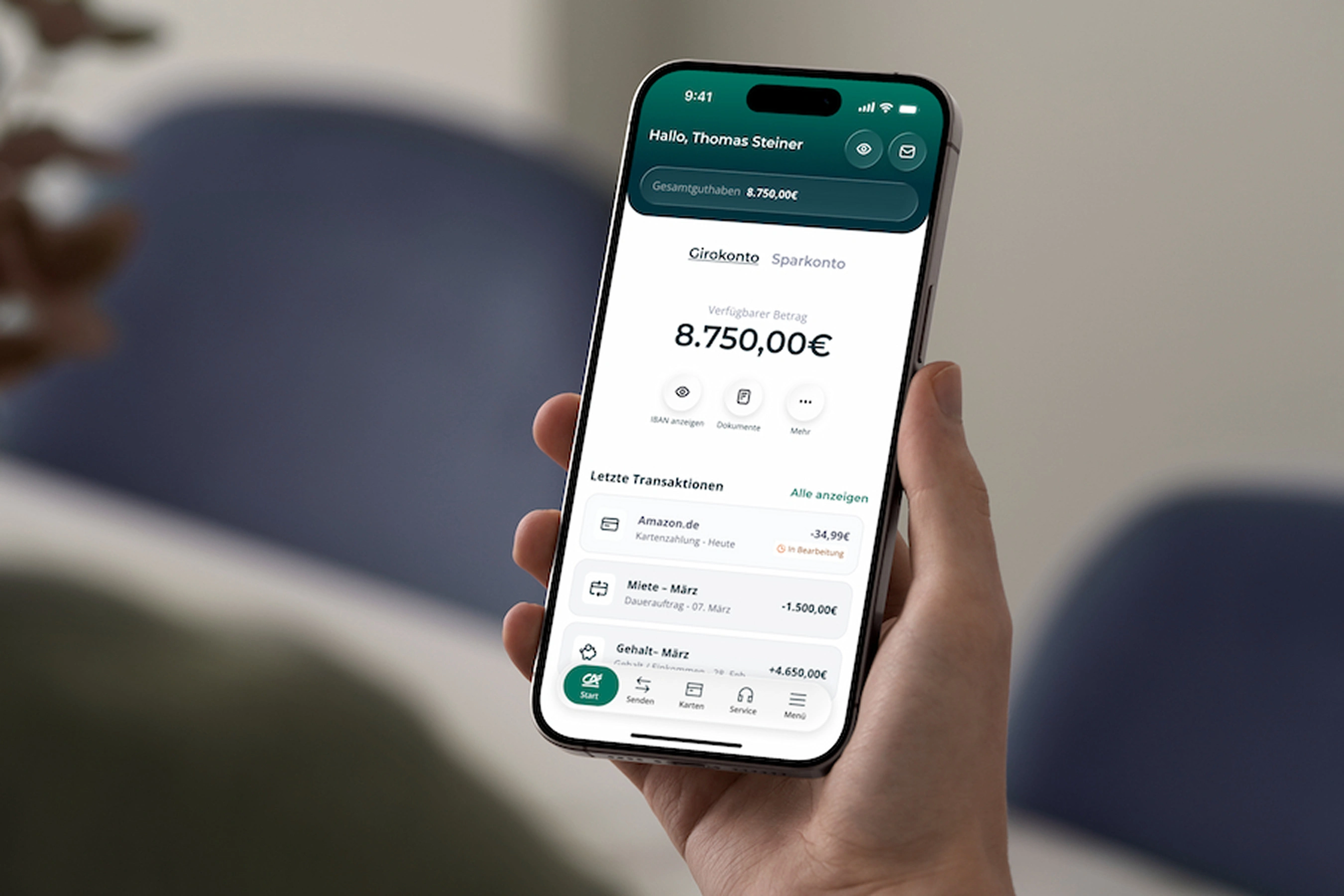 Eine Hand hält ein Smartphone mit geöffneter Banking-App. Auf dem Bildschirm sind der Kontostand und die letzten Transaktionen des Girokontos zu sehen. Im unscharfen Hintergrund ist ein heller Wohnraum mit blauen Sesseln zu erkennen.
