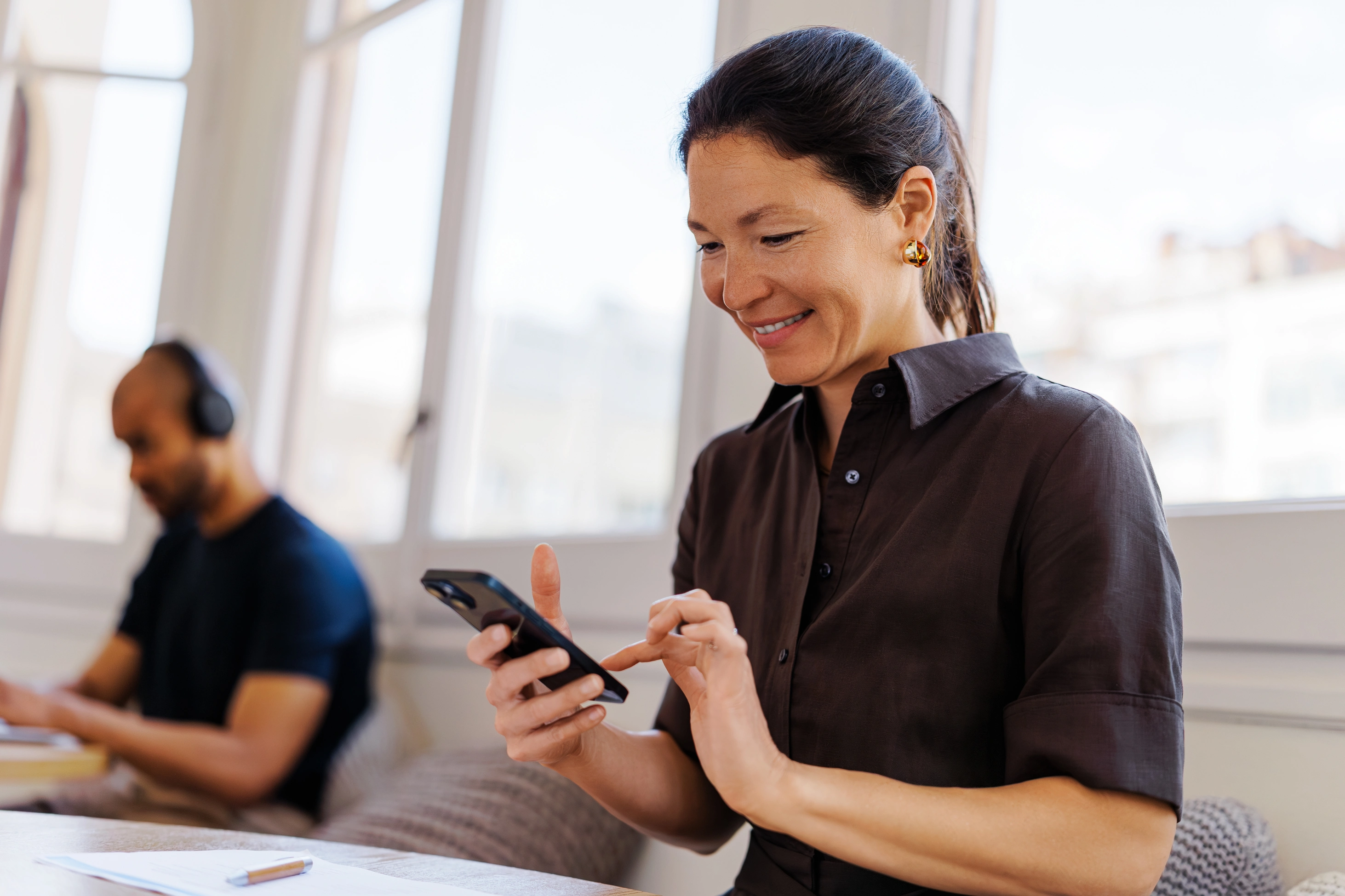 Eine brünette Frau macht an ihrem Arbeitsplatz eine Pause und schaut auf ihr Smartphone.