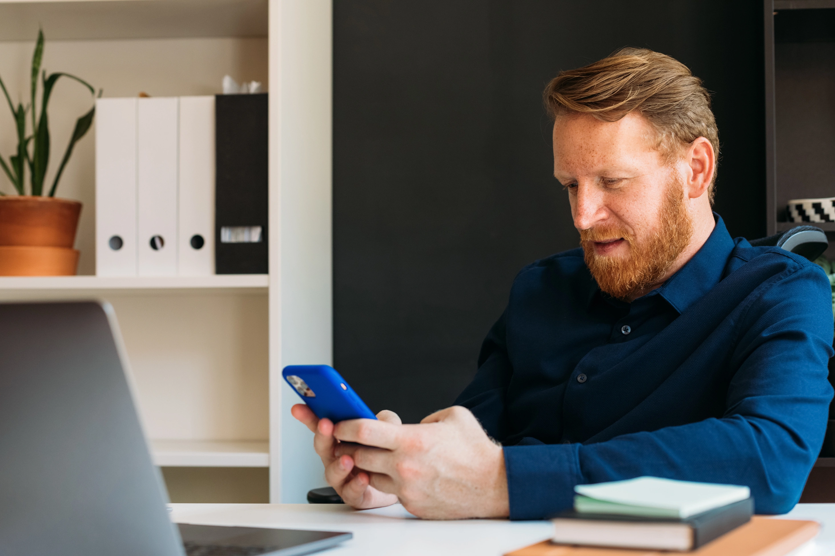 Ein Mann mit rotem Haar und Bart sitzt in einem dunkelblauen Hemd an seinem Arbeitsplatz im Büro und tippt auf seinem Smartphone.