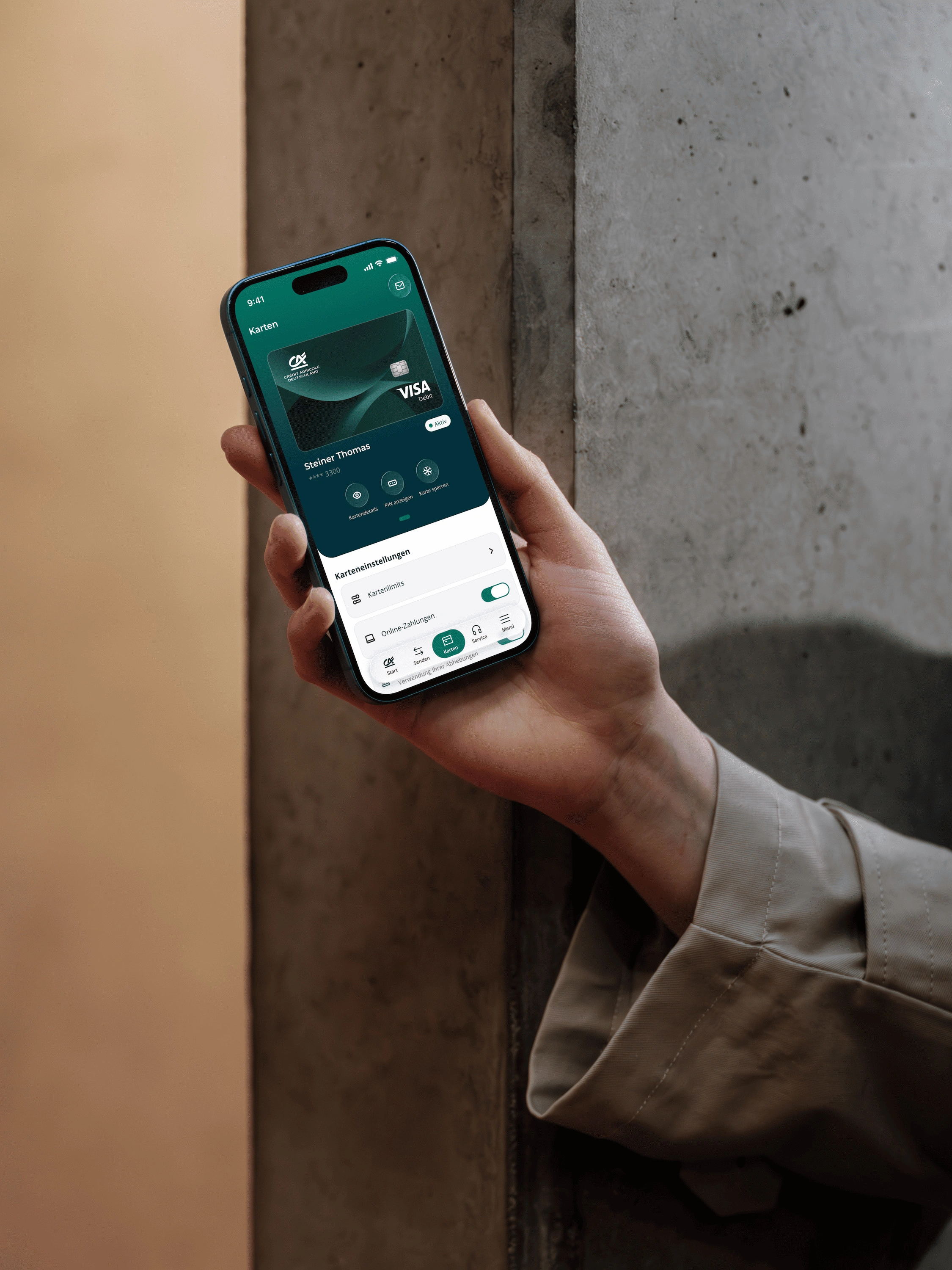 Eine Hand im beigen Trenchcoat-Ärmel hält ein Smartphone mit blauem Rahmen vor eine Betonwand. In der Banking-App ist die Kartenübersicht mit einer Visa-Debitkarte geöffnet, darunter die Karteneinstellungen mit Kartenlimits und Online-Zahlungen.