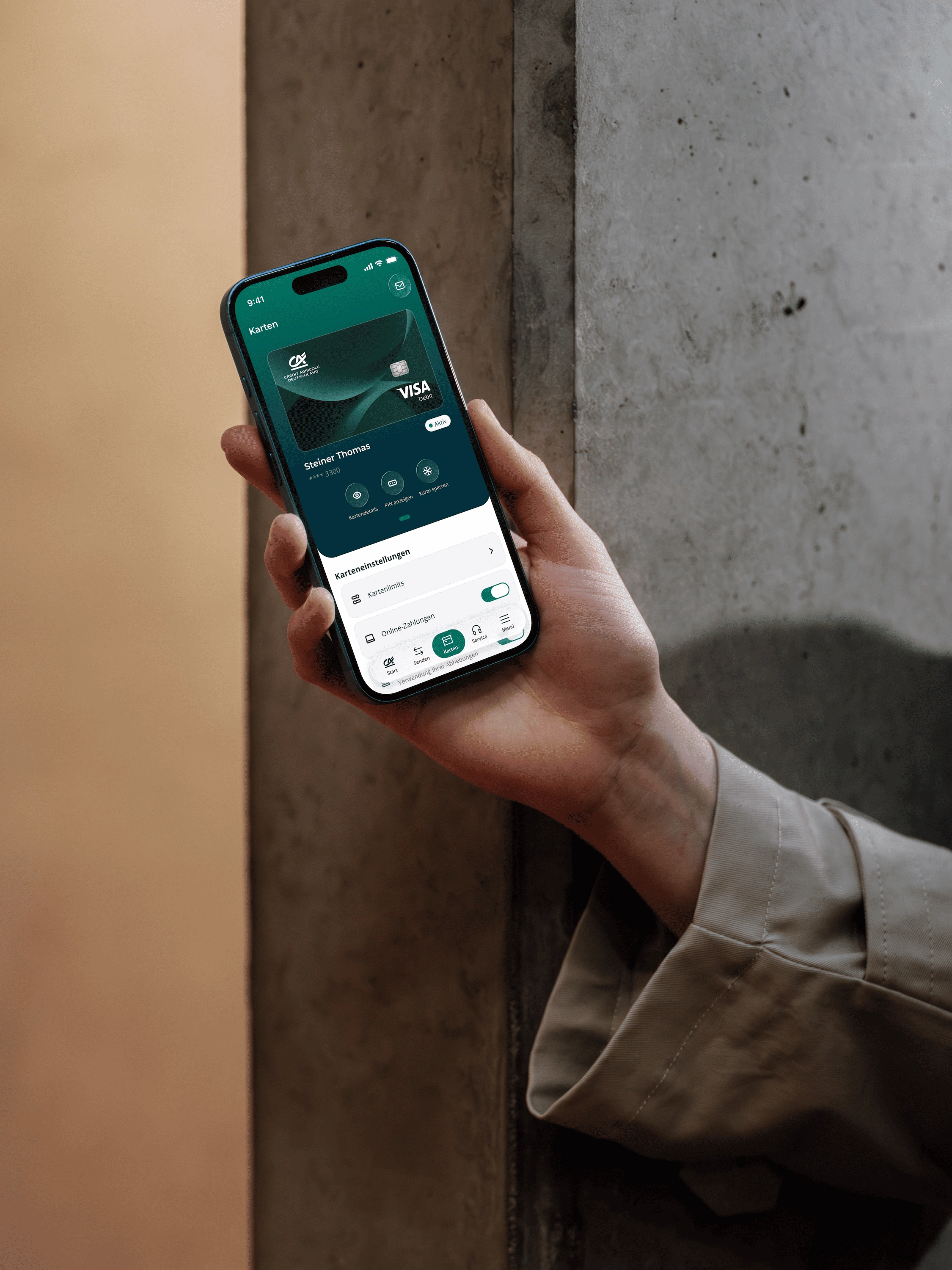 Eine Hand im beigen Trenchcoat-Ärmel hält ein Smartphone mit blauem Rahmen vor eine Betonwand. In der Banking-App ist die Kartenübersicht mit einer Visa-Debitkarte geöffnet, darunter die Karteneinstellungen mit Kartenlimits und Online-Zahlungen.