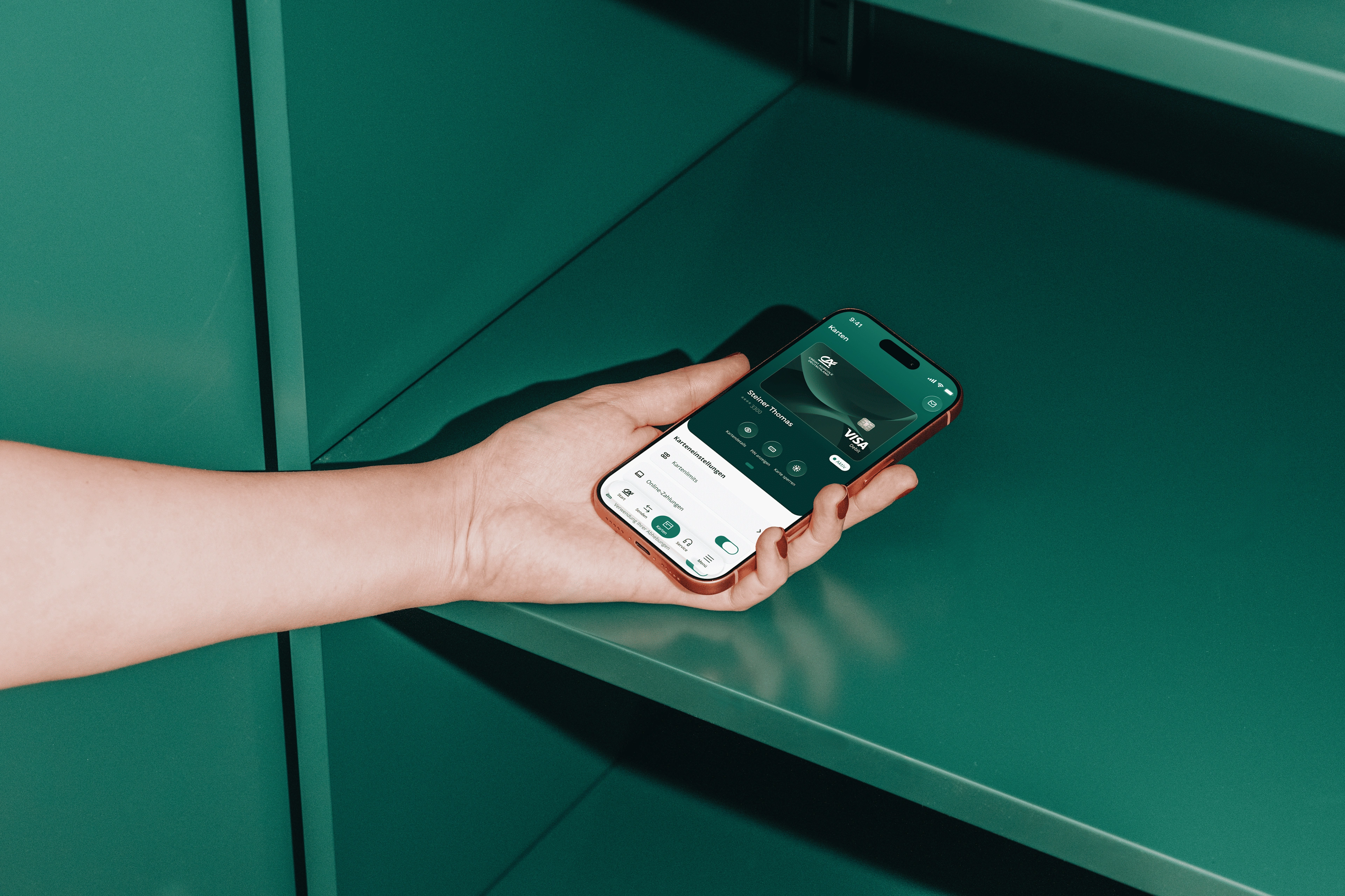 Eine Hand mit rot lackierten Fingernägeln hält ein Smartphone mit orangefarbenem Rahmen vor ein dunkelgrünes Metallregal. In der Banking-App ist die Kartenübersicht mit einer Visa-Debitkarte und den Karteneinstellungen geöffnet.