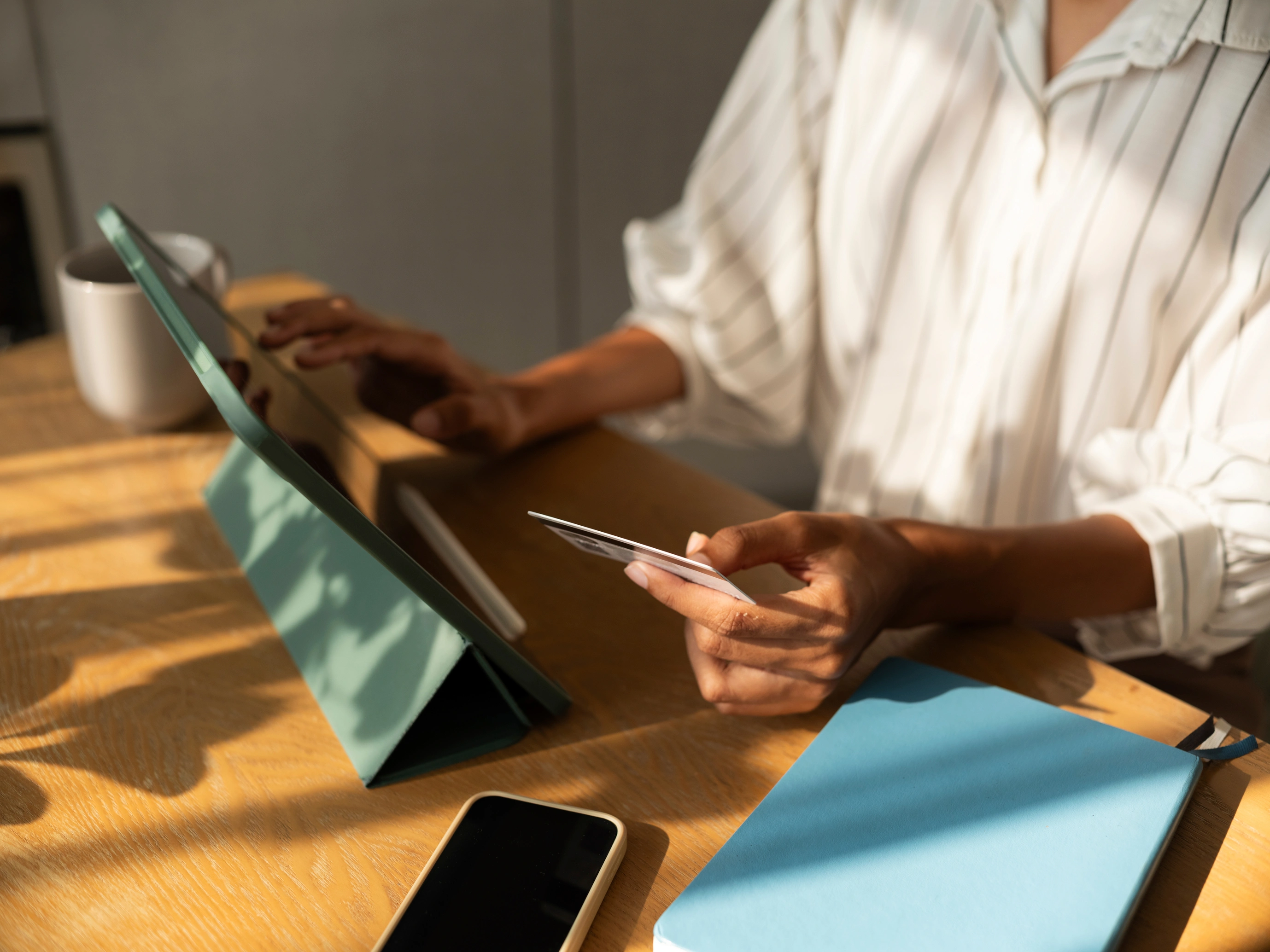 Eine Frau sitzt an einem Tisch und kauft auf ihrem Tablet online ein. In der Hand hält sie eine Kreditkarte. Sonnenlicht fällt auf den Tisch und sorgt für eine gemütliche Atmosphäre.