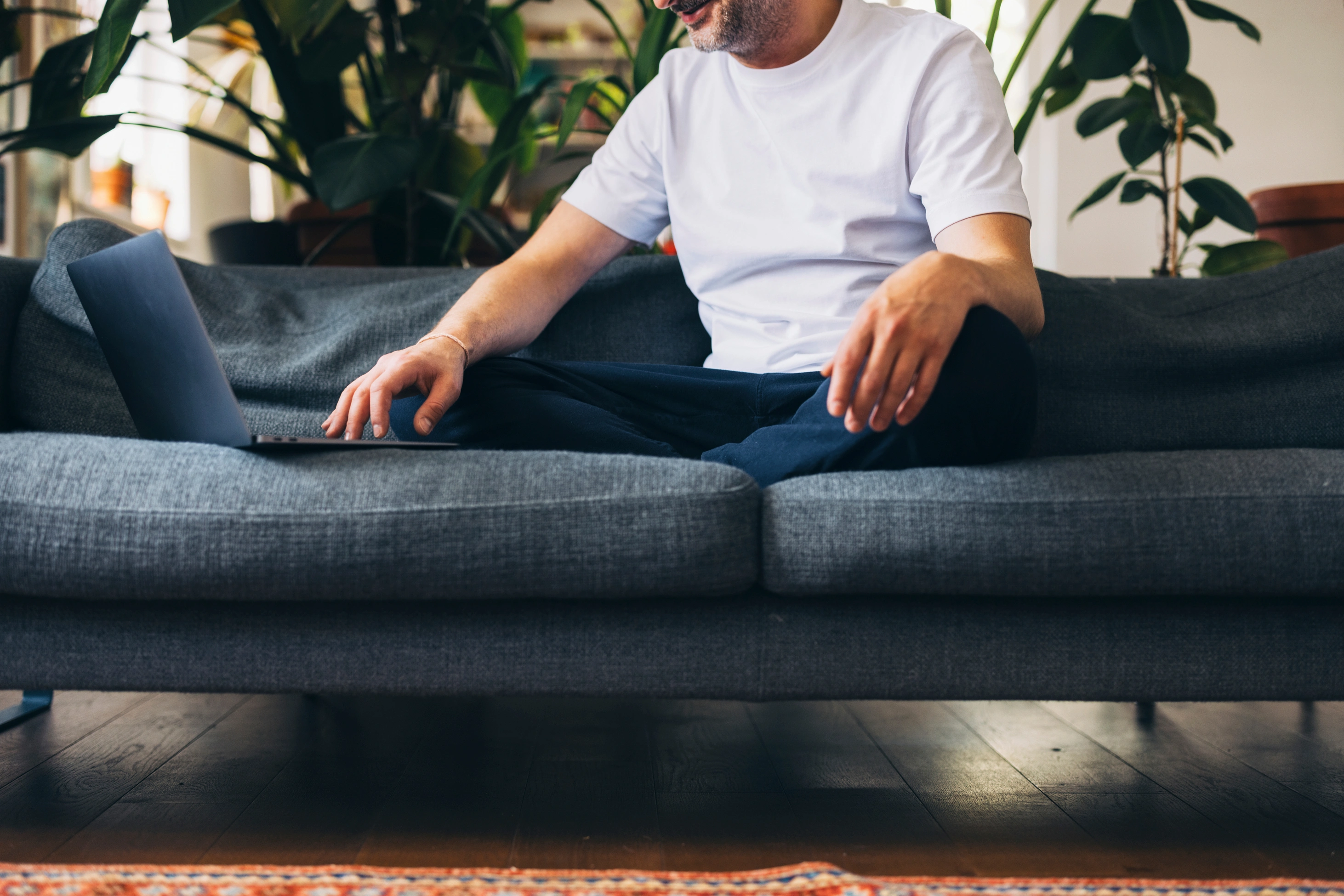 Ein Mann sitzt im Schneidersitz auf einem gemütlichen Sofa und nutzt seinen Laptop, umgeben von Zimmerpflanzen in einer warmen, entspannten Atmosphäre.