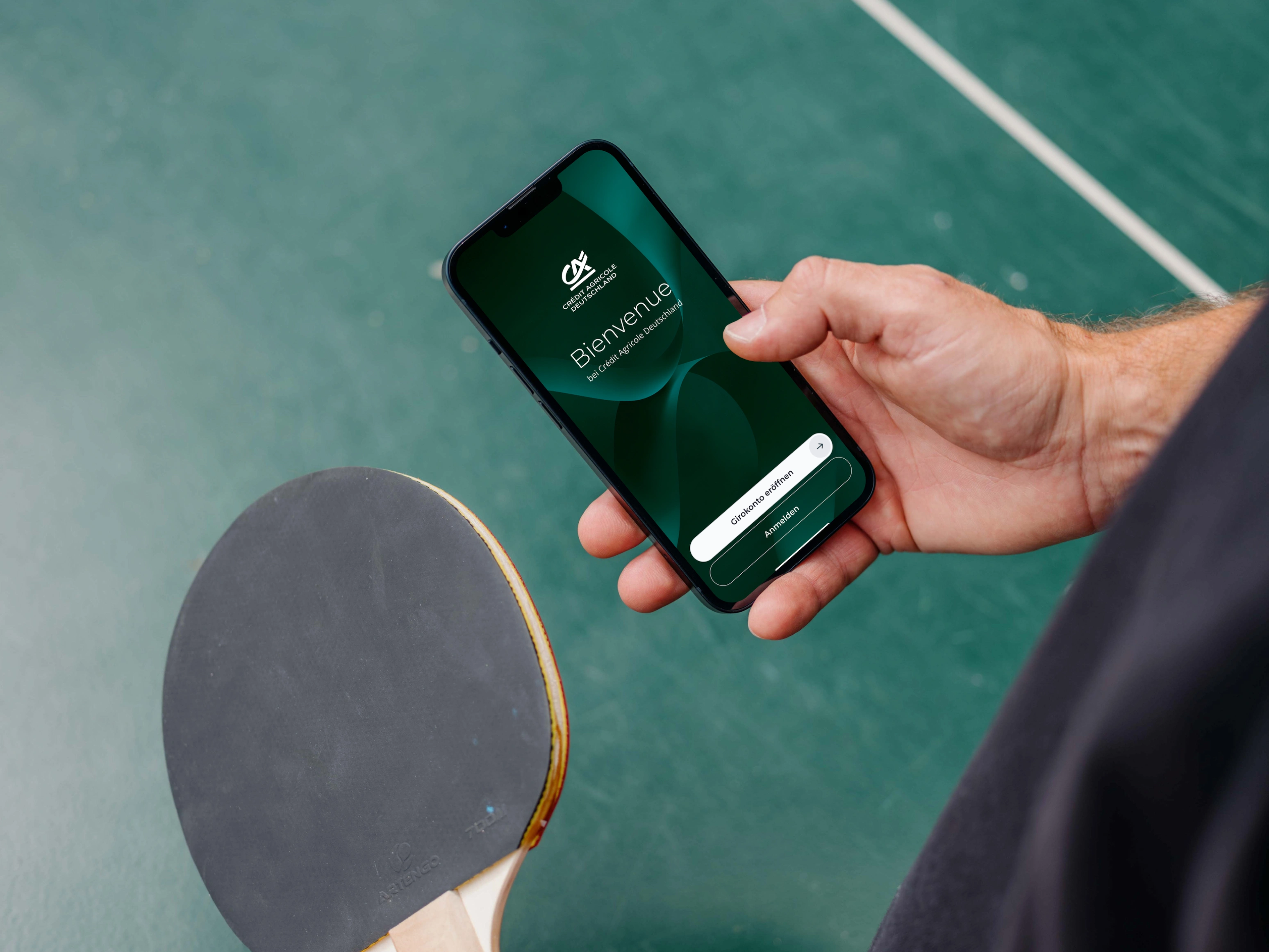 Person hält an einer Tischtennisplatte ein Smartphone mit geöffneter Crédit Agricole Deutschland App in der Hand.