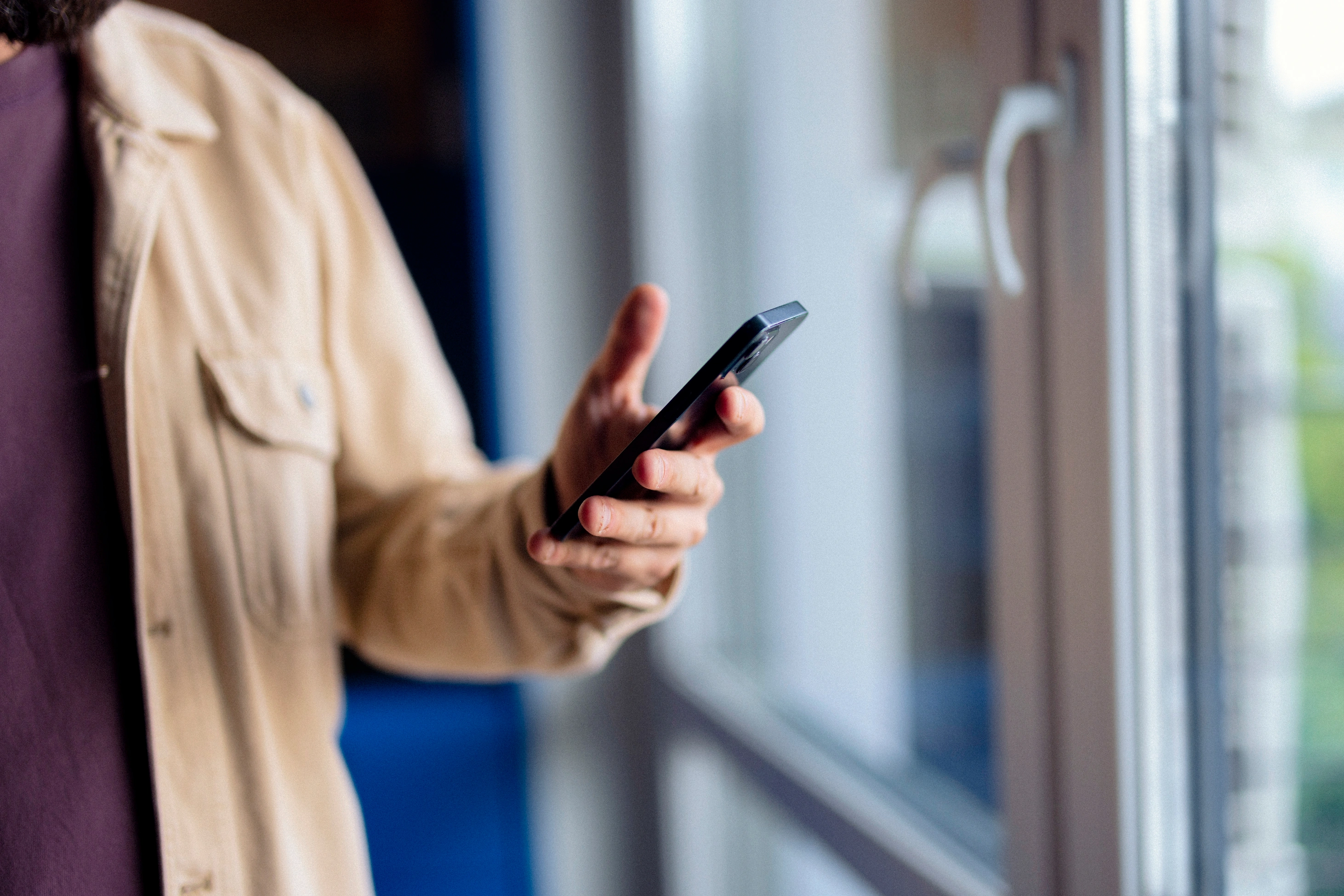 Ein Mann in einer beigen Jacke steht an einem hellen Fenster und hält ein Smartphone in der Hand.