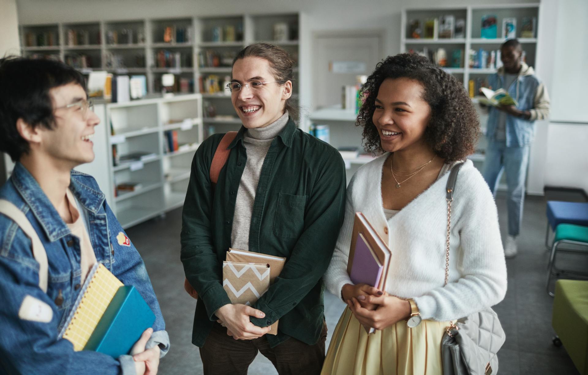 Three students smile and chat in a library, holding books and notebooks. Shelves filled with books line the background. A fourth student is reading in the background. The room is well-lit and welcoming.