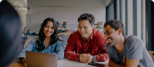 Three people sit at a table, smiling and looking at a laptop screen together in a bright, modern office space with large windows.