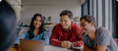 Three people sit at a table, smiling and looking at a laptop screen together in a bright, modern office space with large windows.