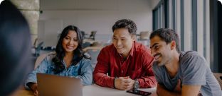 Three people sit at a table, smiling and looking at a laptop screen together in a bright, modern office space with large windows.