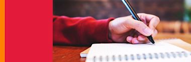 Hand in red sleeve holding black pen over spiral notebook on wooden desk, close-up view of writing