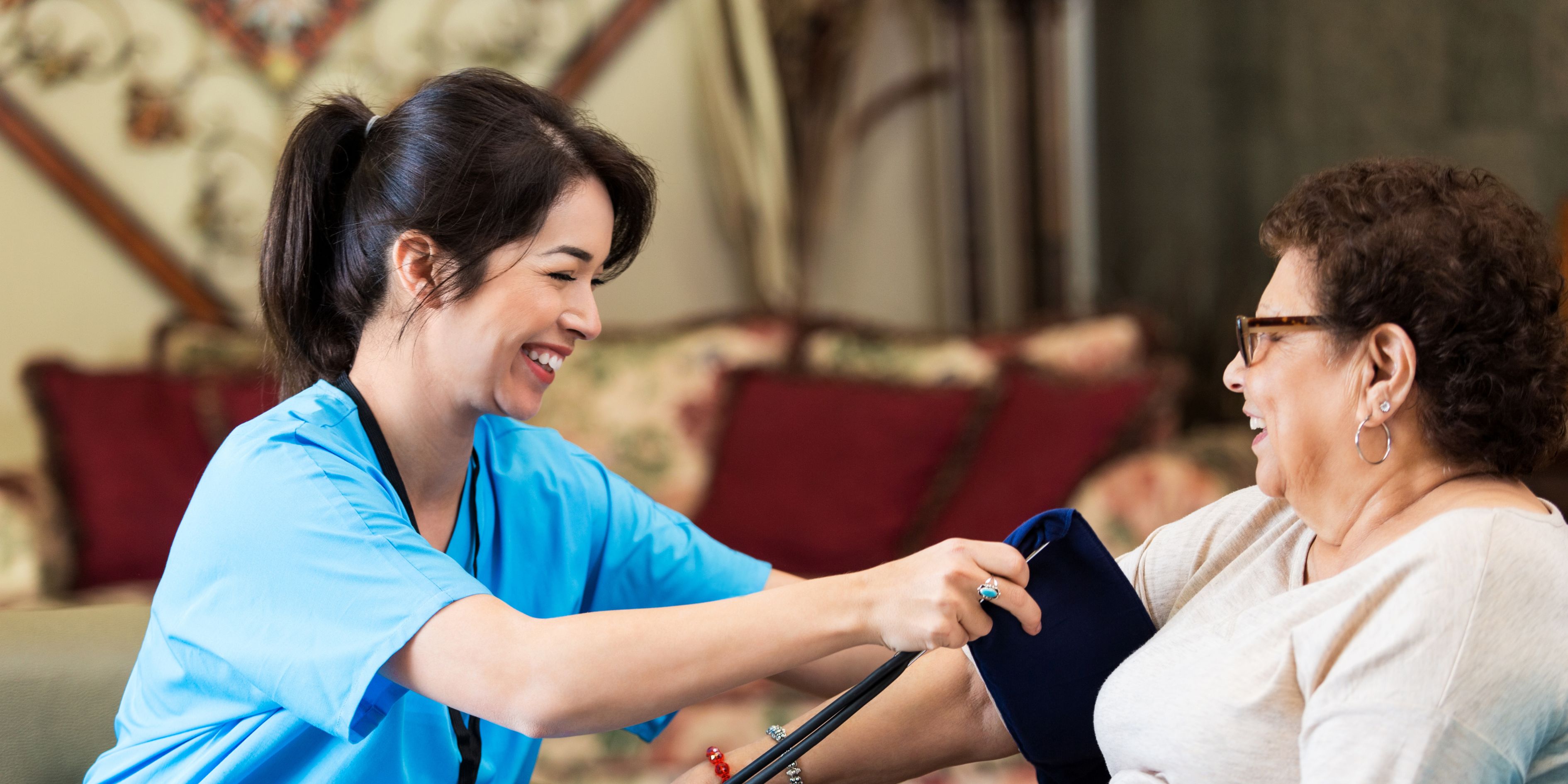 a female nurse, with black hair and fair skin, smiling while she's taking the blood pressure of an elderly woman with curly brown hair and white shirt
