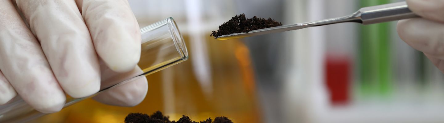 A hand in a lab glove holds a test tube while another hand uses a small spatula to add dark soil. Laboratory equipment and blurred test tubes in the background.