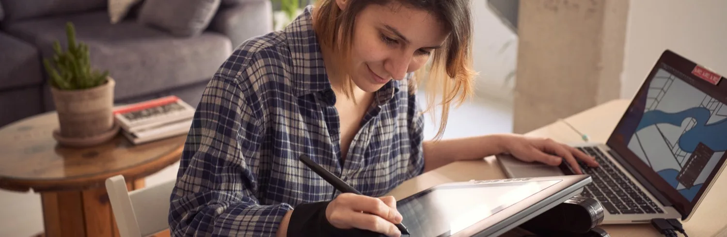 A person wearing a plaid shirt is using a stylus pen on a drawing tablet while looking at a laptop. The setting appears to be an indoor workspace with a potted plant and a couch in the background.