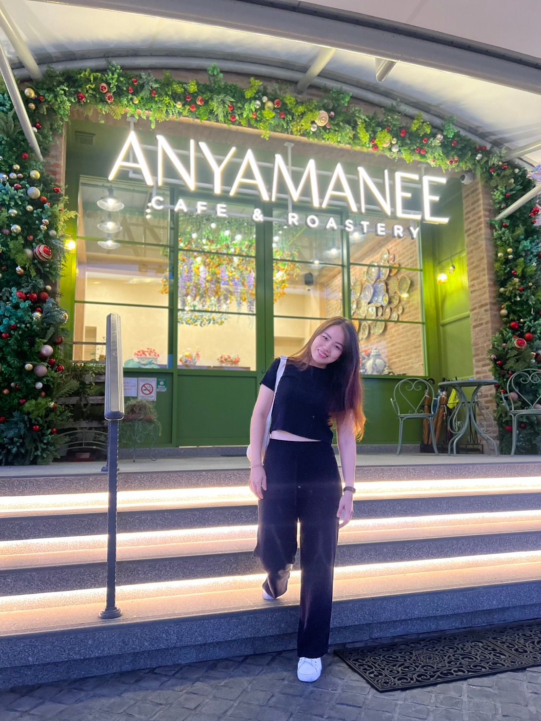 A person smiling and posing on illuminated steps leading to Anyamanee Cafe & Roastery, decorated with festive greenery and ornaments. The modern entrance features glass doors and a welcoming atmosphere.