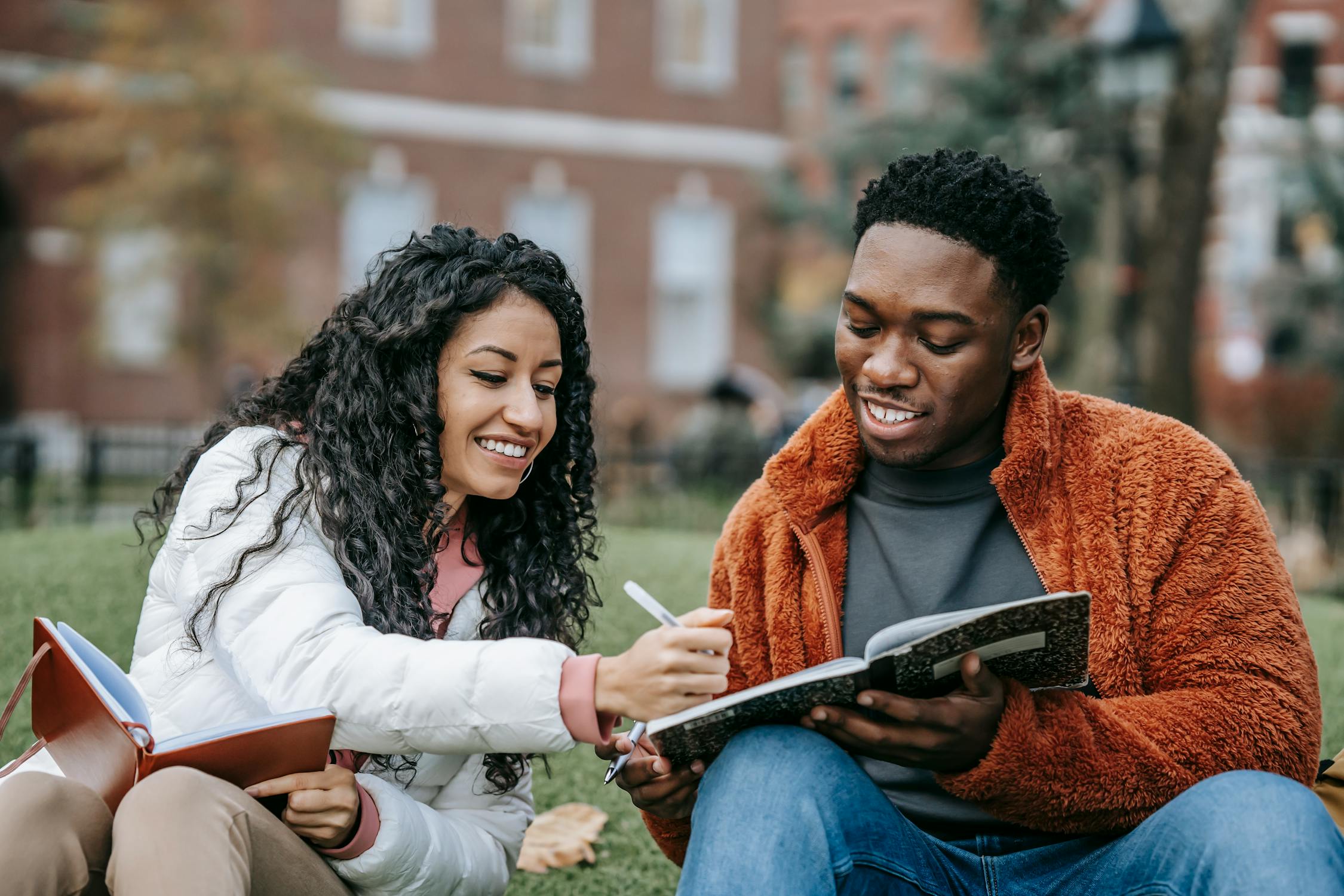 two students outside the university, smiling while looking at each other's notebooks.