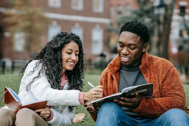 two students outside the university, smiling while looking at each other's notebooks.