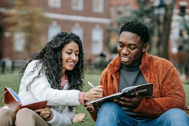 two students outside the university, smiling while looking at each other's notebooks.