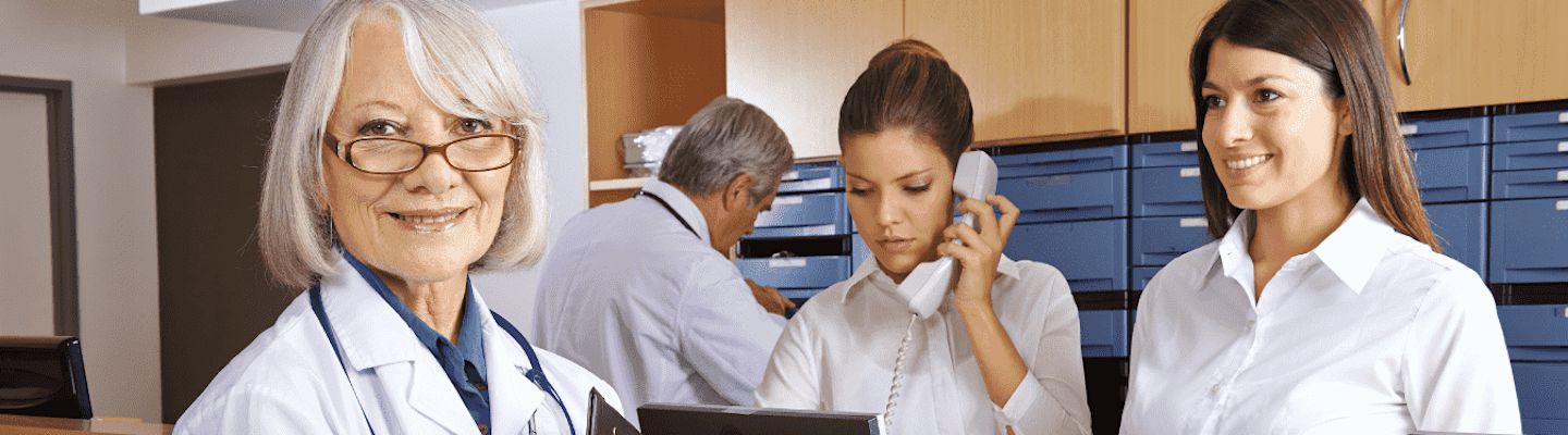 A senior woman in a white coat smiles at the camera, holding a clipboard. Two women in white shirts work behind her; one is on the phone, and the other is looking at the camera. A man in the background is sorting through files.