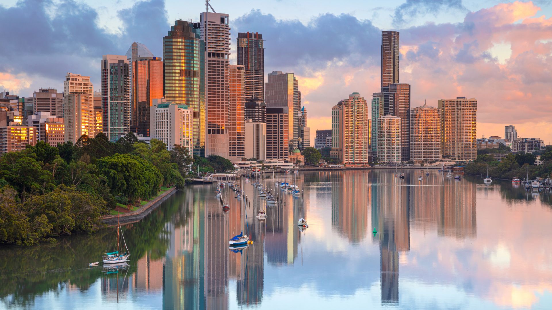 Skyline of a city with tall modern buildings reflecting in a calm river, surrounded by green trees and boats anchored along the water at sunrise or sunset.