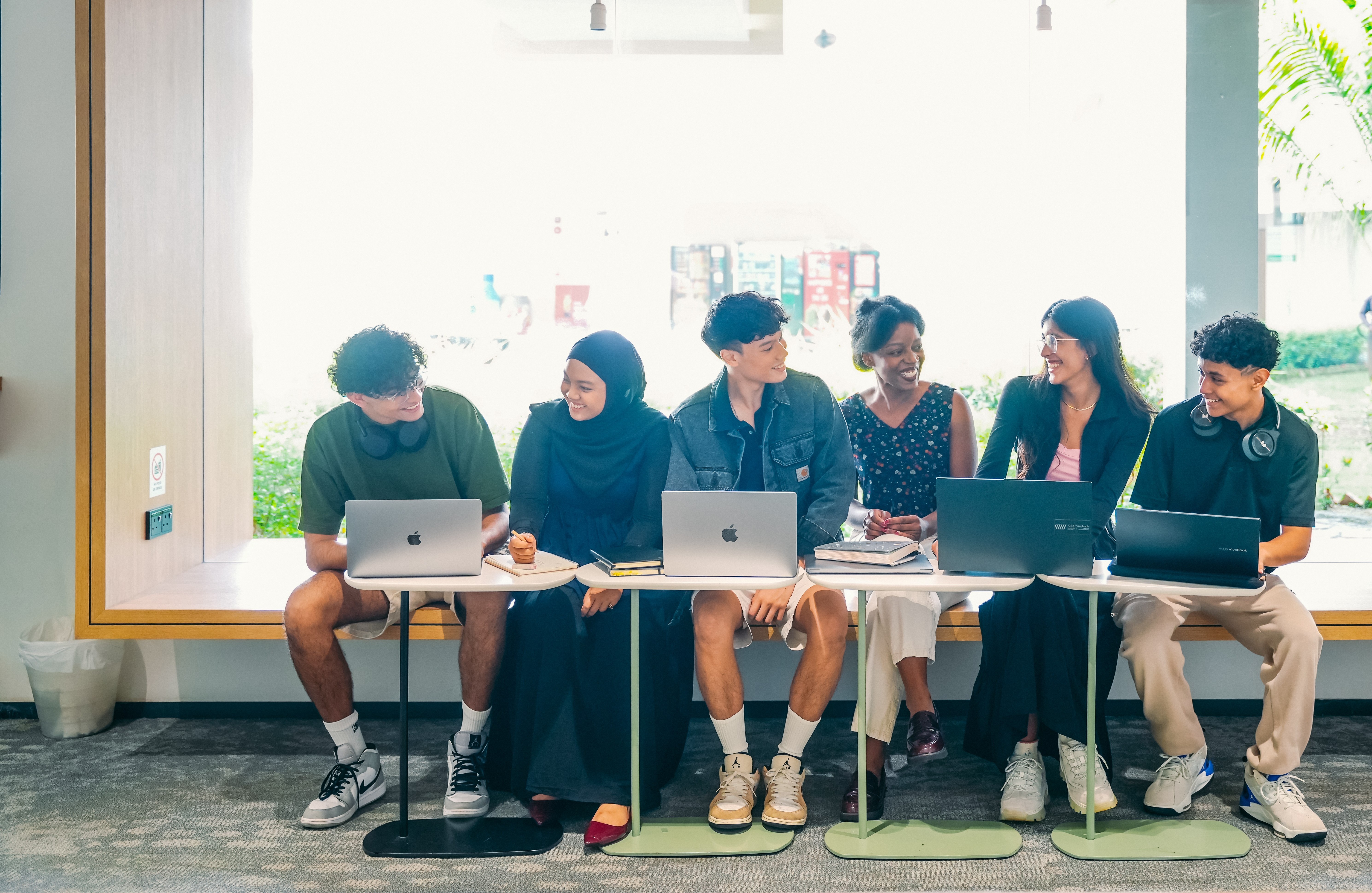Diverse group of students sitting together at study tables with laptops, engaging in conversation and collaborative work in a bright space
