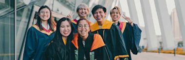  Cheerful graduates in navy and orange academic regalia pose together outside a modern building, making peace signs and smiling