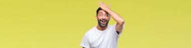 A man with a beard and wearing a white t-shirt stands in front of a yellow background, smiling and touching his forehead as if he just remembered something.