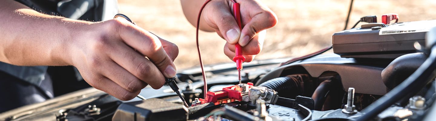 Hands using a multimeter to test a car battery, with jumper cables attached and engine components visible in background