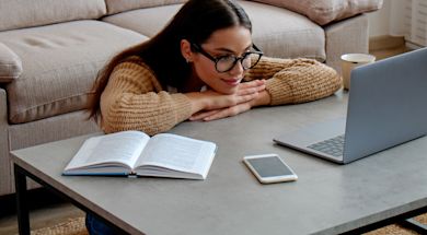 A woman with glasses, wearing a brown sweater, leans on a table with a laptop, an open book, and a smartphone. She appears relaxed, with a sofa and a cup in the background.