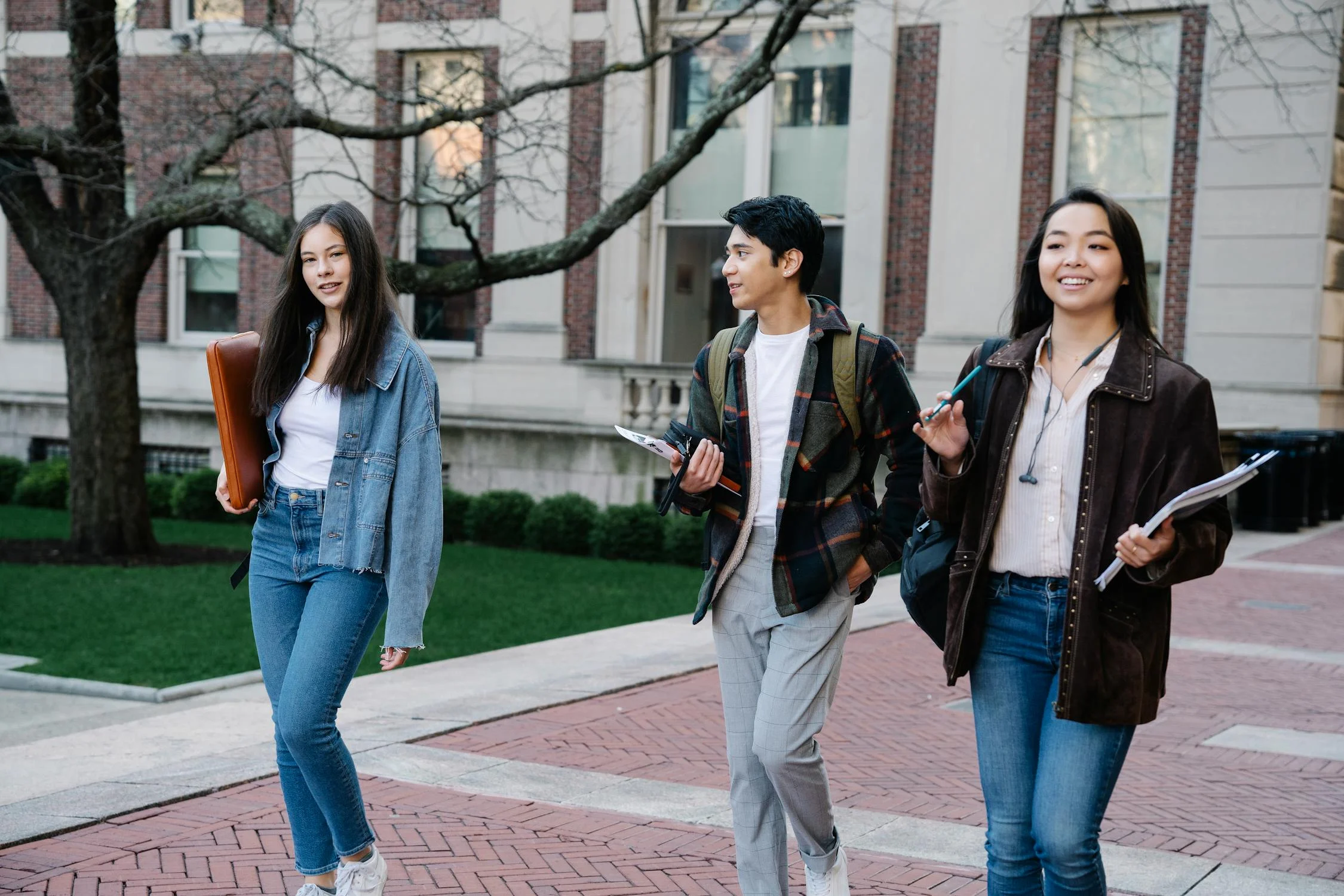 Three young adults walk on a campus path, holding books and notebooks. Theyre casually dressed in jeans and jackets, with a building and lawn in the background. One person looks towards the others, who are smiling.