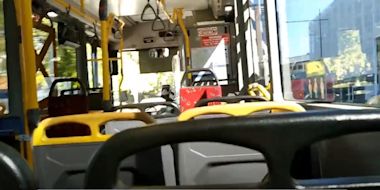 Interior view of a public bus showing empty seats, yellow handrails, and windows with sunlight streaming through