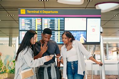 Một nhóm ba sinh viên đang đứng trong nhà ga và bàn về các quy định thị thực mới trên điện thoại của họ.