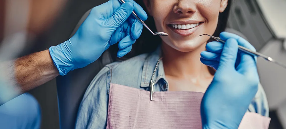 A person wearing a pink dental bib smiles while a dentist, wearing blue gloves, examines their teeth using dental instruments.