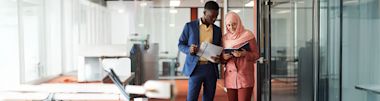 Two professionals, one wearing a blue suit and the other in a pink suit and hijab, stand in a modern office hallway, reviewing documents together.