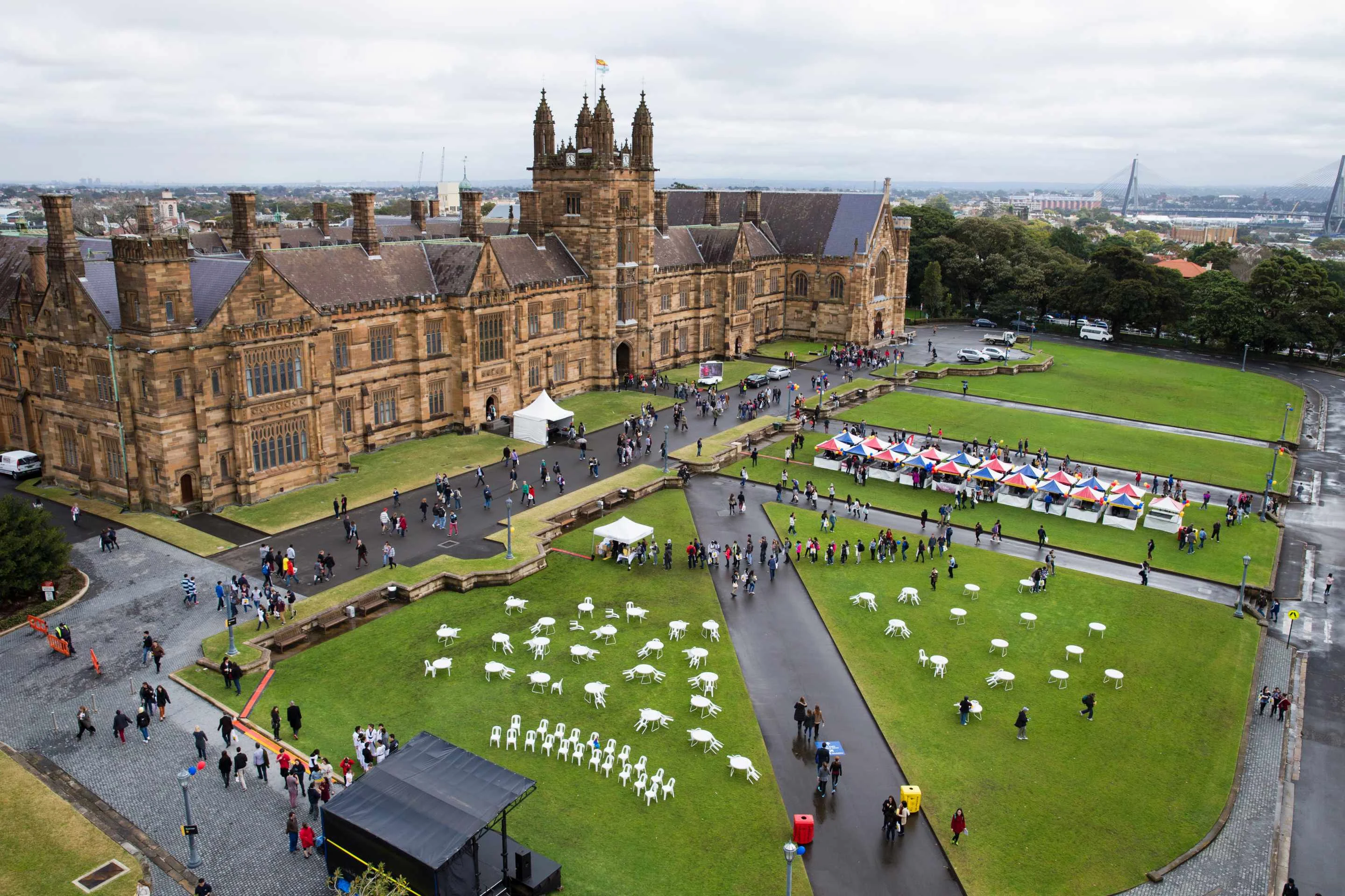 Aerial view of University of Sydney's Gothic sandstone buildings with crowds and white chairs scattered on lawns during an outdoor event