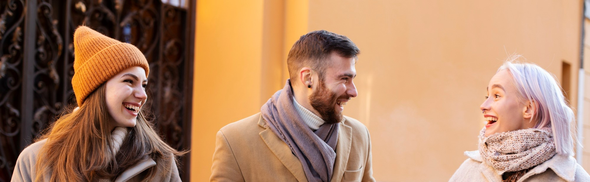 Three people in winter clothing stand outside a building, laughing. A woman in a brown beanie, a man with a beard and scarf, and a woman with white hair in a knitted scarf enjoy the moment.