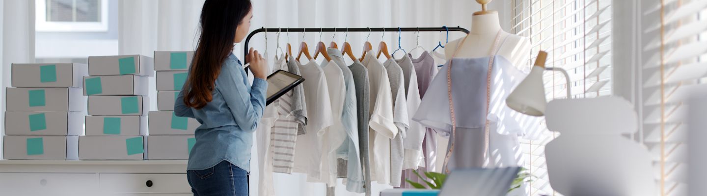 A woman in a blue shirt inspects clothes on a rack with a tablet in hand. The room is filled with stacked boxes, and a mannequin displays a light blue dress. White blinds cover the windows, casting soft light into the room.