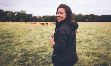 A person in a black jacket stands smiling in an open field with a herd of deer grazing in the background. The scene is set under a cloudy sky with trees lining the horizon.