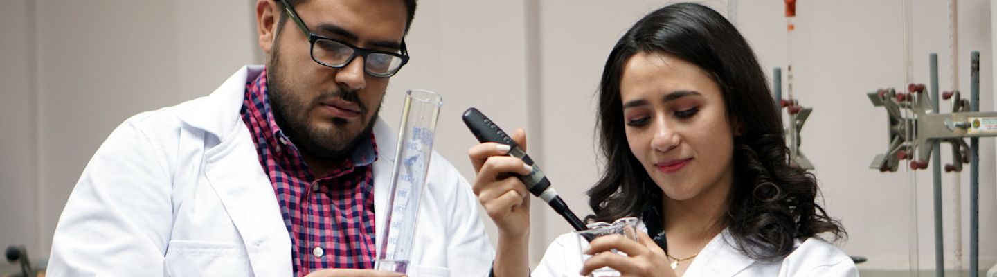 Two scientists in white lab coats examine laboratory equipment and test tubes while conducting research in a laboratory setting.